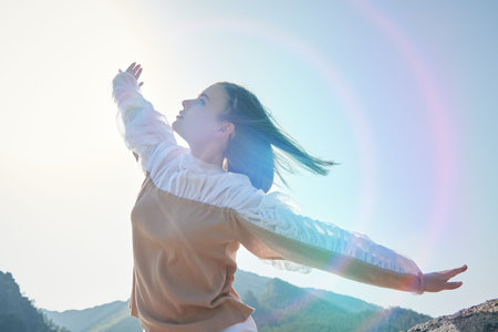 Adorable girl in a blouse with wide sleeves stands on top of a mountain with outstretched arms...