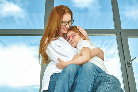 Mother and her little daughter happily smile and hug sitting by the window in apartment. Happy family.の写真素材