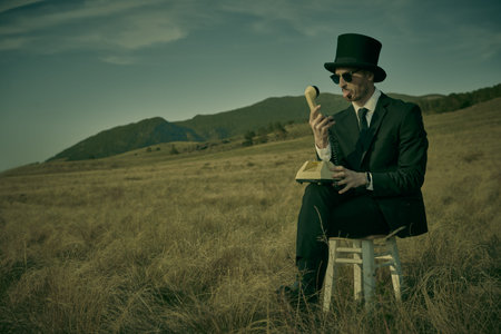 Surrealism and subreality. A gentleman in a black suit and top hat sits on a stool in a field and...