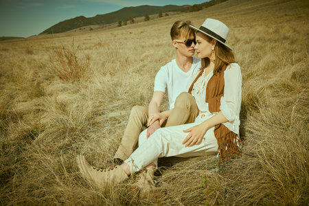 Romantic young couple in stylish summer clothes sitting close to each other on the dry grass in the...