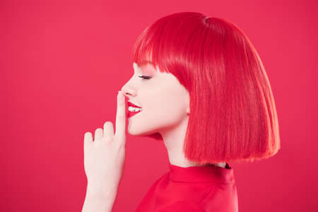 Sideview portrait of a pretty, smiling girl with bright red hair holding her index finger to her red lips. Red background. Beauty, makeup. Hairstyle, hair coloring. Fashionable red colour.の写真素材