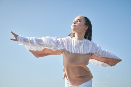 Female beauty and fashion. Adorable girl model in fashionable blouse with wide sleeves stands on the background of a blue sky enjoying warm rays of sun and warm wind. Travel and vacation.の写真素材