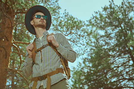 Tourism, nature exploring. Portrait of a handsome young man backpacker standing in a pine forest and looking into the distance. Active rest, hiking, adventure.の写真素材
