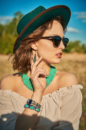 An attractive boho girl in a feminine blouse, hat and sunglasses on the background of a field and blue sky. Summer vacation, traveling. Bohemian, modern hippie style.の写真素材