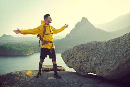 Feeling of freedom, achievement. A man tourist with a backpack behind his back stands on top of a mountain with outstretched arms, enjoying the scenery around. Active lifestyle, hiking.の写真素材