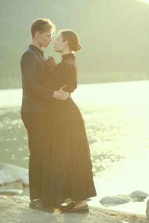 Elegantly dressed loving couple stand close to each other by the sea at sunset tenderly looking at...