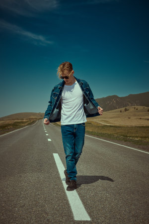 Portrait of a young man hitchhiker in denim clothes and black sunglasses walking on the highway on...