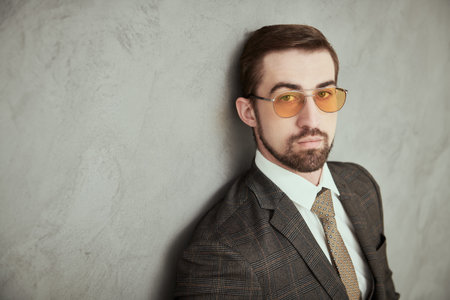 Men's fashion. Portrait of a handsome man in elegant suit, tie and sunglasses posing by a gray wall.の写真素材