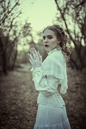 Historical reconstruction of the Victorian era. Portrait of a beautiful noble lady in a white dress walking in an old park. Hairstyle and makeup, late 19th century. Fashion.の写真素材