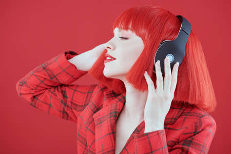 DJ, party girl. A stylish girl with bright red lips and red hair wearing a red checkered jacket and headphones. Red studio background. Beauty, Fashion.の写真素材