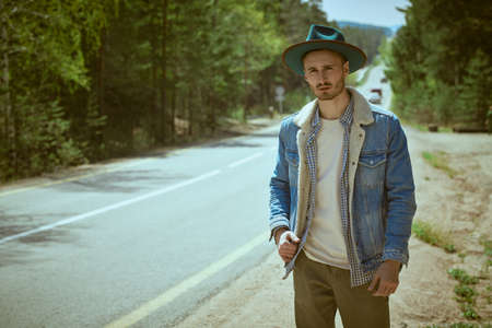 Handsome hipster man in denim clothes and hat stands by the road waiting for a passing car. Road travel hitchhiking. Denim fashion.の写真素材