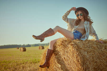 Full length portrait of a romantic girl sitting on a haystack in a field in the rays of the setting sun. Modern hippie and bohemian style. Summer and autumn fashion.の写真素材