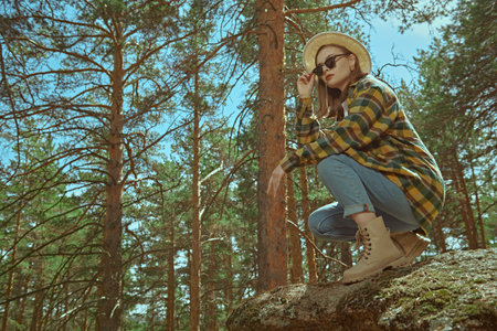 Full length portrait of a hipster girl in jeans, a plaid shirt and a hat, standing in a pine forest. Summer vacation in nature. Tourism. Denim fashion.の写真素材