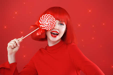 Beautiful joyful girl with bright red lips and red shoulder-length hair poses with a lollipop. Red studio background with shining lights. Beauty, cosmetics, makeup. Hairstyle, hair coloring. Holiday.の写真素材