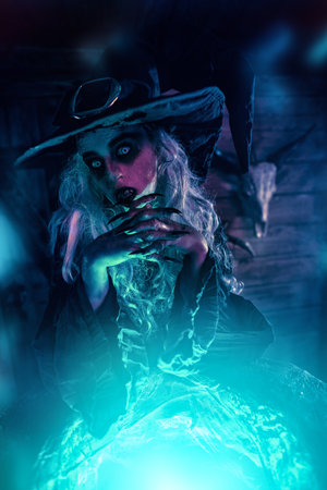 A terrible evil witch stands leaning on her cauldron, illuminated by magical blue light. Halloween. Scary stories, fantasy.の写真素材