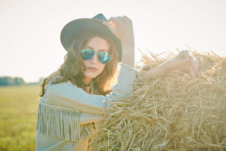 Modern hippie and wild west style. Beautiful fashion model girl poses in denim clothes, a hat and sunglasses by a haystack in a field in the rays of the setting sun. Summer and autumn fashion.の写真素材