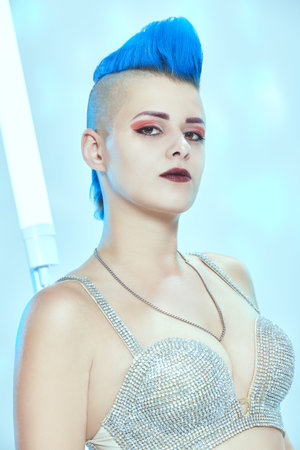 Portrait of a bright girl punk rock musician with bright makeup and blue mohawk posing at studio...