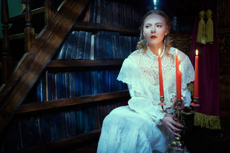 Historical mystical novel. A beautiful lady aristocrat in a white lace dress sits in a dark vintage library by candlelight looking warily away. Victorian style.の写真素材