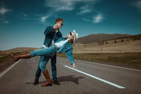 Portrait of a beautiful couple in denim clothes and black sunglasses dancing on a highway in a hilly valley. Freedom and independence. Road adventures, travelling. Denim style.の写真素材