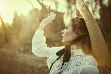 Portrait of a beautiful romantic girl in a white dress and a straw hat walking in an old autumn park at sunset. Victorian style.の写真素材