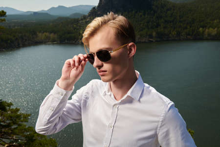 A purposeful young man in a white shirt and black sunglasses stands against a lake and mountain landscape. Business concept. Vacation and travel.の写真素材