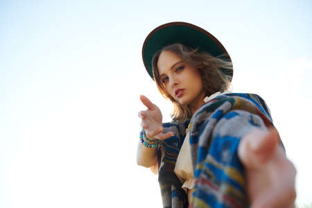 A modern girl in boho style clothes and hat reaches out her hand to the camera in the background of the sky. Bohemian, hippie style. Summer mood. Vacation and travel. Copy space.の写真素材