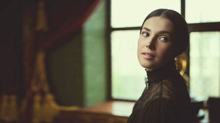 An aristocratic girl with black, gathered hair, and in a black strict dress, sits in a room with...