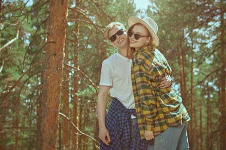 Happy hipster couple of travelers stand in a sunny pine forest joyfully hugging and smiling. Summer vacation outdoors. Tourism. Denim fashion.の写真素材