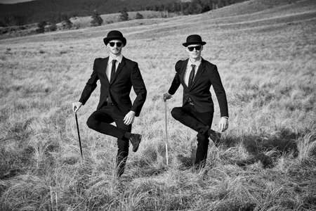 Two well-groomed gentlemen in black suits, bowler hats and black sunglasses dance synchronously with walking sticks in an open wild field. Surrealist style. Black and white shot.の写真素材