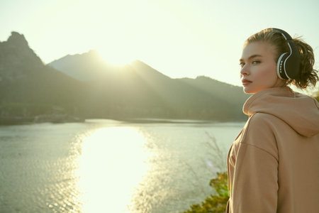 A happy girl listens to pleasant music on headphones at sunset against a beautiful sea landscape....
