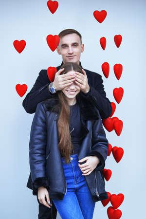 Valentine's day. Handsome young man closes the eyes of a joyful, smiling girl. Happy young people in love. White background with hearts decorations. Love and relationship.の写真素材