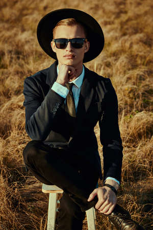 Fashion, business style. Portrait of a handsome blond man in an elegant black suit, white shirt, a hat and black sunglasses sitting on a stool in sun-scorched field.の写真素材