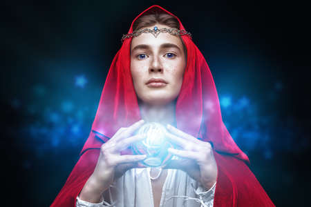 A young prince of elves in a red velvet cloak and a crown on his head holds a magic shining sphere in his hands. Studio portrait on a black background. Fairy tale, magic. Fantasy.の写真素材