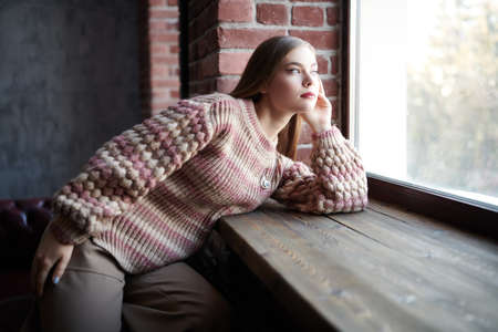 A lovely fair-haired girl looks dreamily out the window in a loft style room. People and emotions.の写真素材