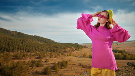 Bright summer style in clothes and accessories. Stylish girl poses in bright clothes and pink sunglasses on top of a mountain against a valley and sky background. High fashion. Copy space.の写真素材