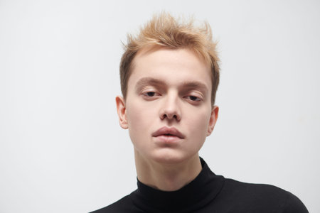 Studio shot of a young man against a white wall in a black turtleneck looking calmly at the camera....