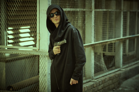 A dangerous teenage boy dressed in a black hooded cardigan and black sunglasses stands on a back street near a metal mesh fence with a gun in his hand. Juvenile delinquency. Difficult teenagers.の写真素材
