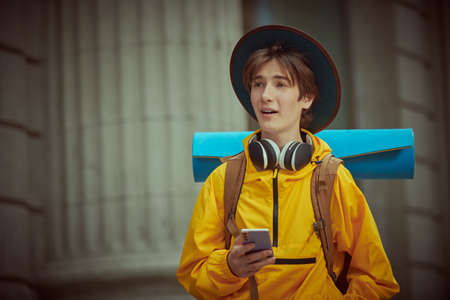A young tourist, a teenager boy with a backpack and a tourist rug behind his shoulders, is sightseeing the city with a smartphone in his hand. Holidays, travel. Active, educational lifestyle.の写真素材
