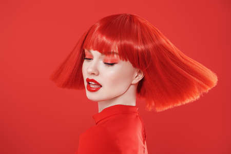 Adorable girl with bright red lips and red hair. Red studio background. Beauty, makeup. Hairstyle, hair coloring. Fashionable red colour.の写真素材