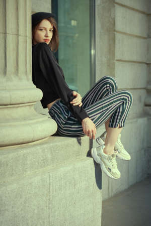 A carefree pretty girl with a bob haircut dressed in a black beret, a black blouse and striped trousers sits by the column on a city street. Style and Fashion. Summer mood.の写真素材