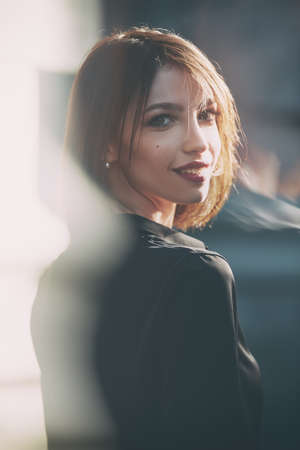 A lovely smiling young woman with a bob haircut in a black blouse stands on a city street looking back and smiling charmingly.の写真素材