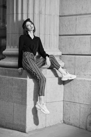 Style and Fashion. A pretty brunette girl with a bob haircut dressed in a black blouse and striped trousers sits by the column of the building on city street. People, lifestyle. Black-and-white shot.の写真素材