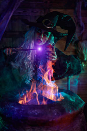 Halloween. Scary tales. Horrible old witch with a magic wand in her hands brews witchcraft in her cauldron illuminated by magical light.の写真素材