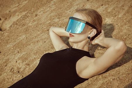 Perfect slim blonde girl lies on the sand in a black swimsuit and mirrored sunglasses. Desert landscape. Fashion and futuristic style.の写真素材