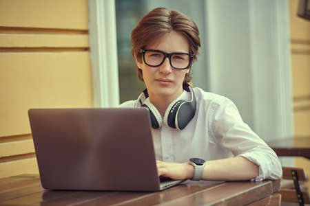 A teen boy in a white shirt and glasses sits at an outdoor cafe table with his laptop. Modern generation. Education, student portrait.の写真素材