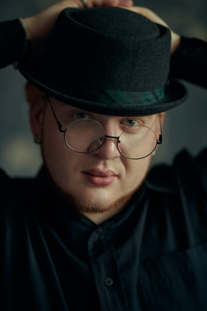 A red-haired young man with chubby cheeks wearing a black shirt, stylish black hat and round glasses...