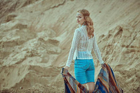 A beautiful girl in boho style clothes and jewelry stands in a desert among sand dunes holding an...