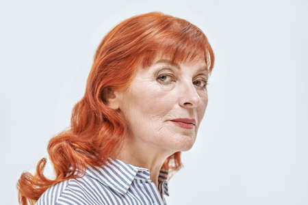 Senior people. A beautiful elderly woman with bright red hair wearing a striped shirt. Studio portrait on a white background. Beauty, make-up and cosmetics for the elderly.の写真素材