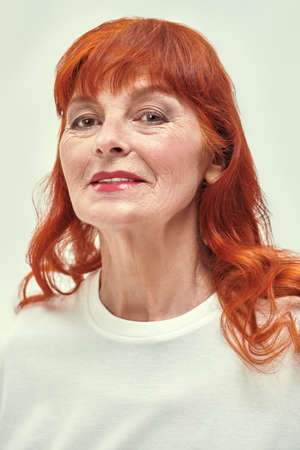Studio shot of a happy beautiful elderly woman with bright red hair and elegant makeup wearing a white blouse. White background. Senior people concept. Make-up and cosmetics for the elderly.の写真素材
