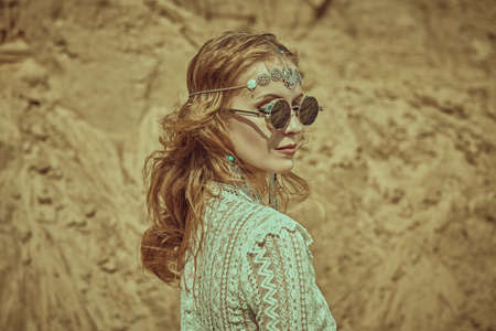 A lovely blonde woman in elegant boho style jewelry and clothes stands against the backdrop of sand dunes and smiles. Summer bohemian style. Fashionable shot.の写真素材